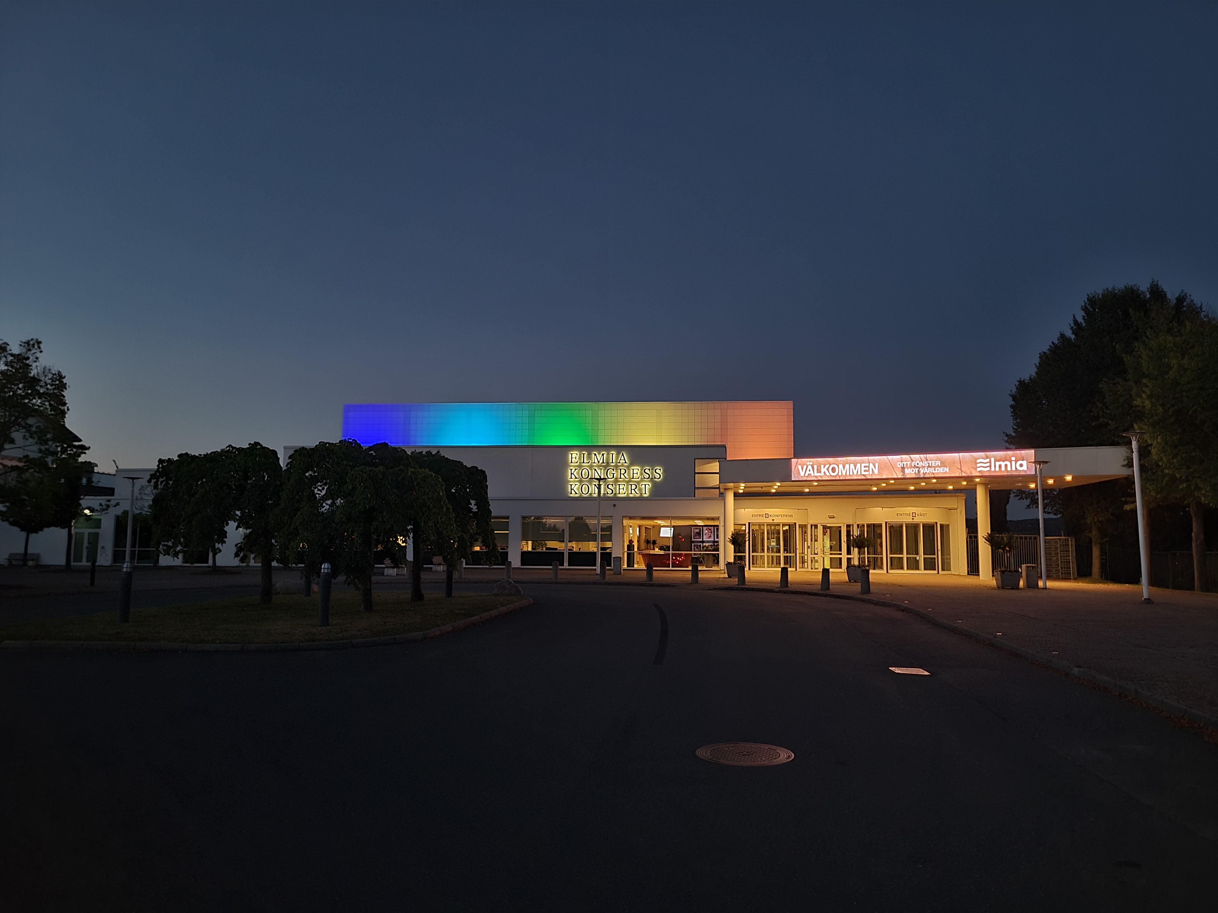 Elmia Kongress och Konserthus upplyst i regnbågsfärger, sett från parkeringen mot entrén.